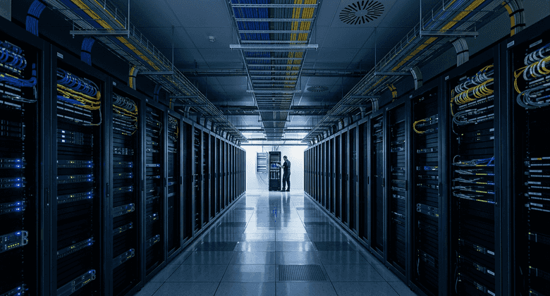 A network engineer works at a patch panel at the end of a server rack aisle, cable bundles running overhead under blue status lights.